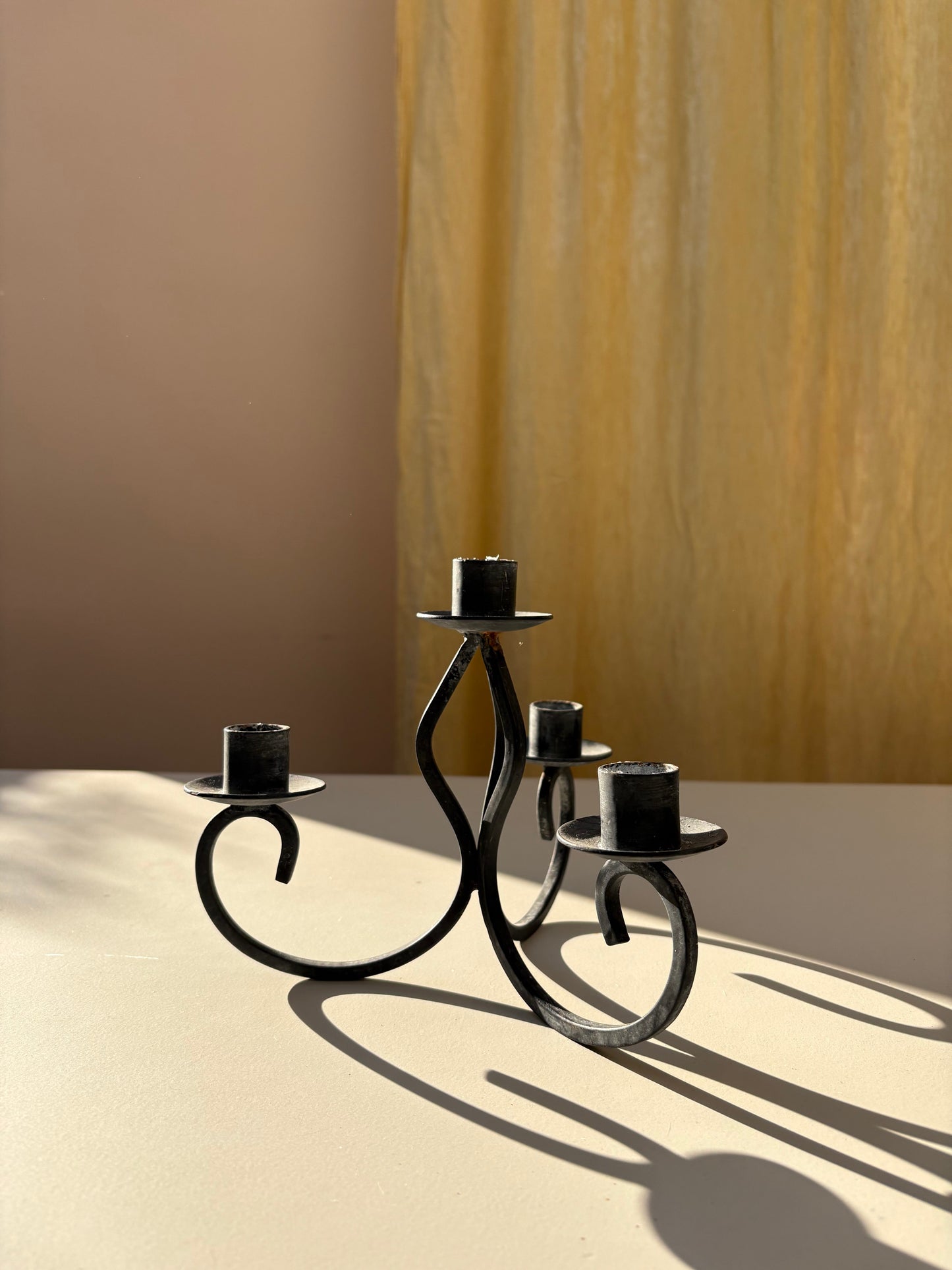 A black wrought iron five-arm candelabra candle holder with a sculptural scroll design, displayed on a beige surface with dramatic sunlight and shadows against a split beige and gold background.